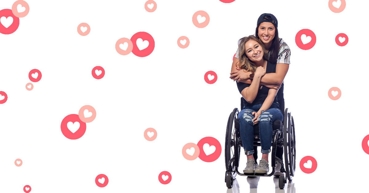 A young, 22-year-old brunette woman wearing dark ripped jeans and a black shirt with white vans sits in her wheelchair. Another dark-haired woman wearing a black backwards hat and also wearing a white, blue and brown striped shirt, stands behind her, hugging her from behind.