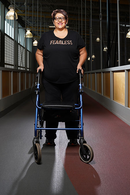 Teresa Begay confidently stands holding the handles of her walker on a running track. She smiles at the camera.