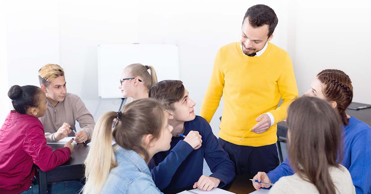 A teacher stands near a table where youth are sitting. They are all talking.