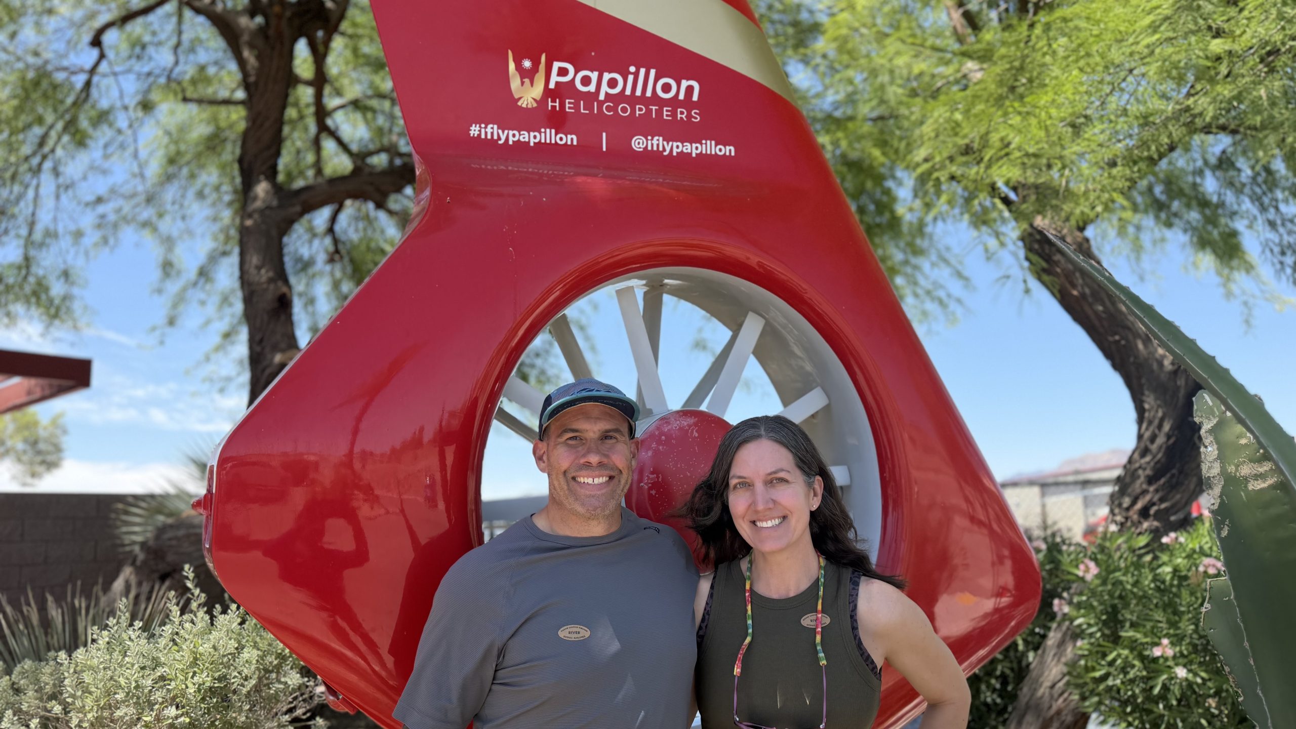 A man wearing a hat and a woman wearing a green tank top pose for a photo in front of a red helicopter statue. 