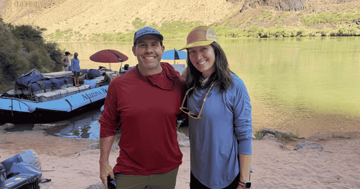 Current image: A man and a woman are pictured in the foreground in front of a river and a blue raft with two people standing in it. The pair is ready to volunteer.