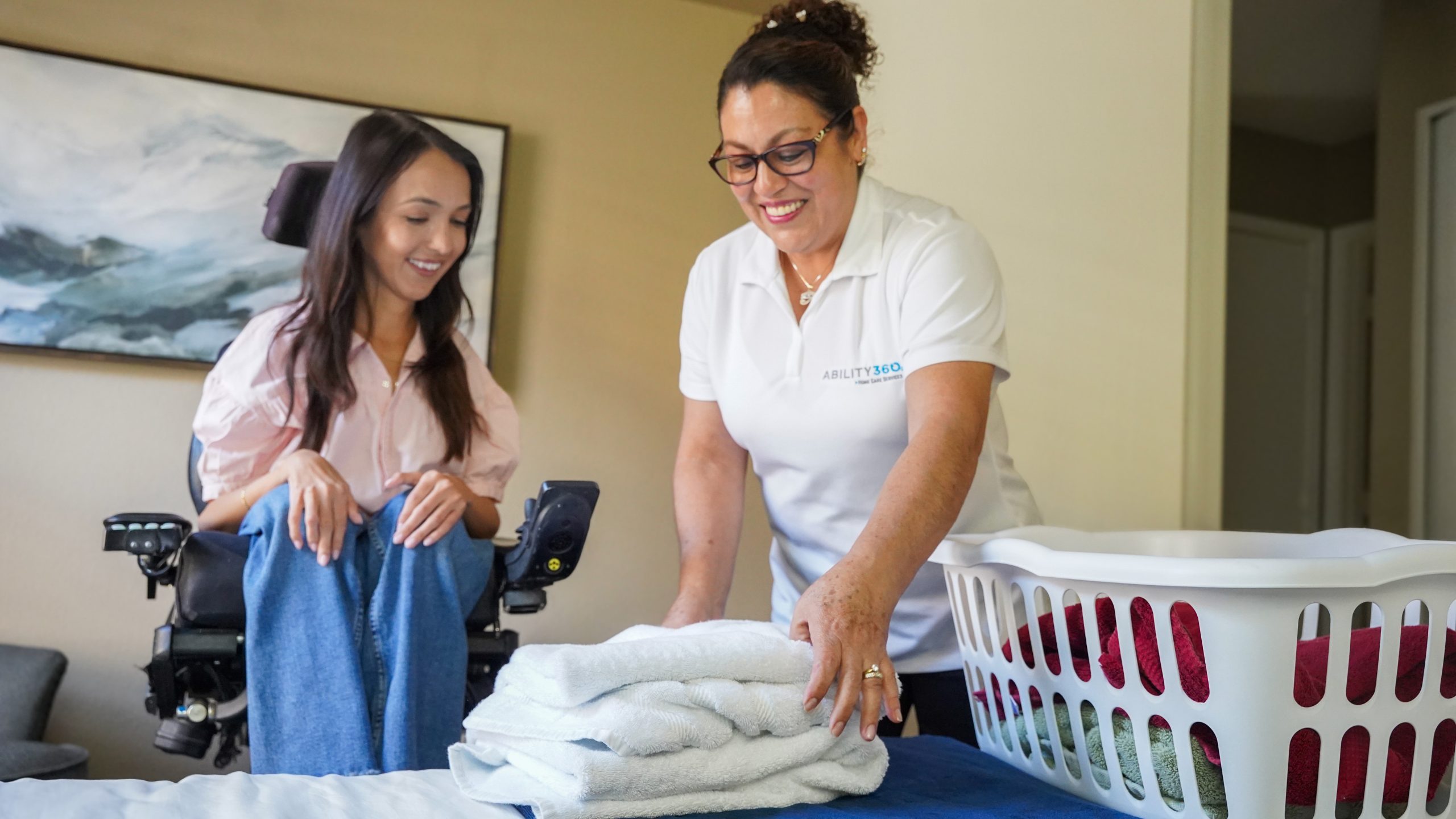 A in-home care attendant folds laundry alongside a care recipient who is sitting in a power wheelchair.