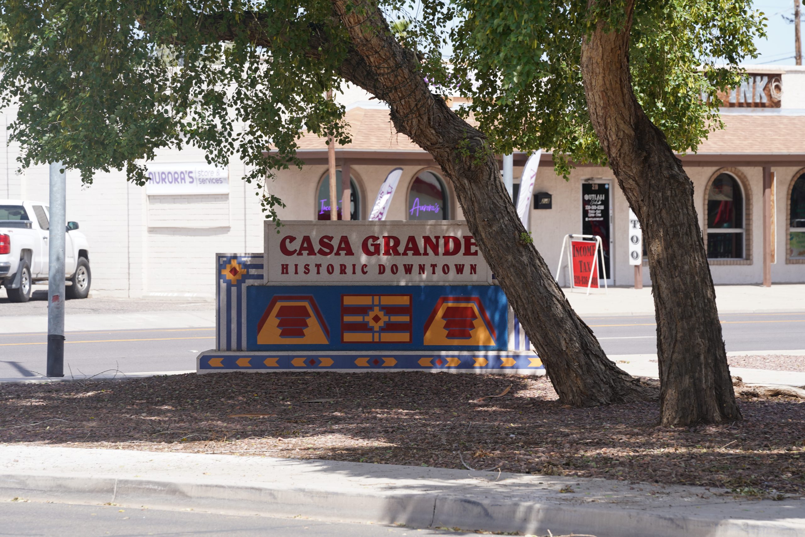 a photo of the downtown casa grande sign. it sits in the shade of a large tree.